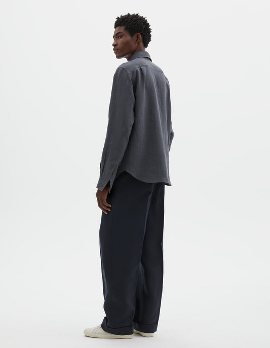 Full body image, back view of model wearing dark grey cotton long sleeve shirt, with mother of pearl button cuffs and pleats on back yoke. Styled with dark navy blue trousers and off white canvas trainers.