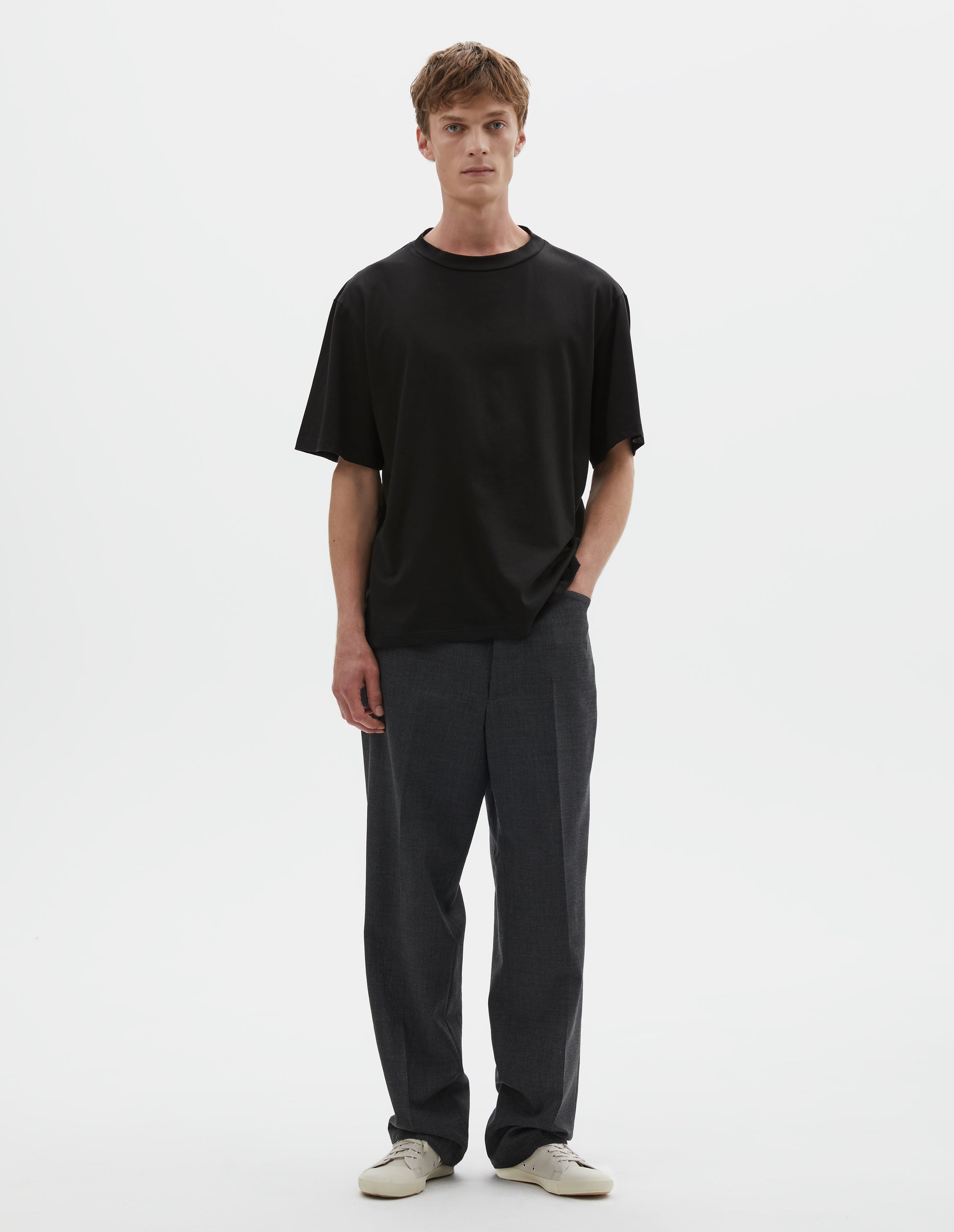 Full body image, front view of model wearing black cotton short sleeve t-shirt. Styled with grey trousers and beige canvas trainers.