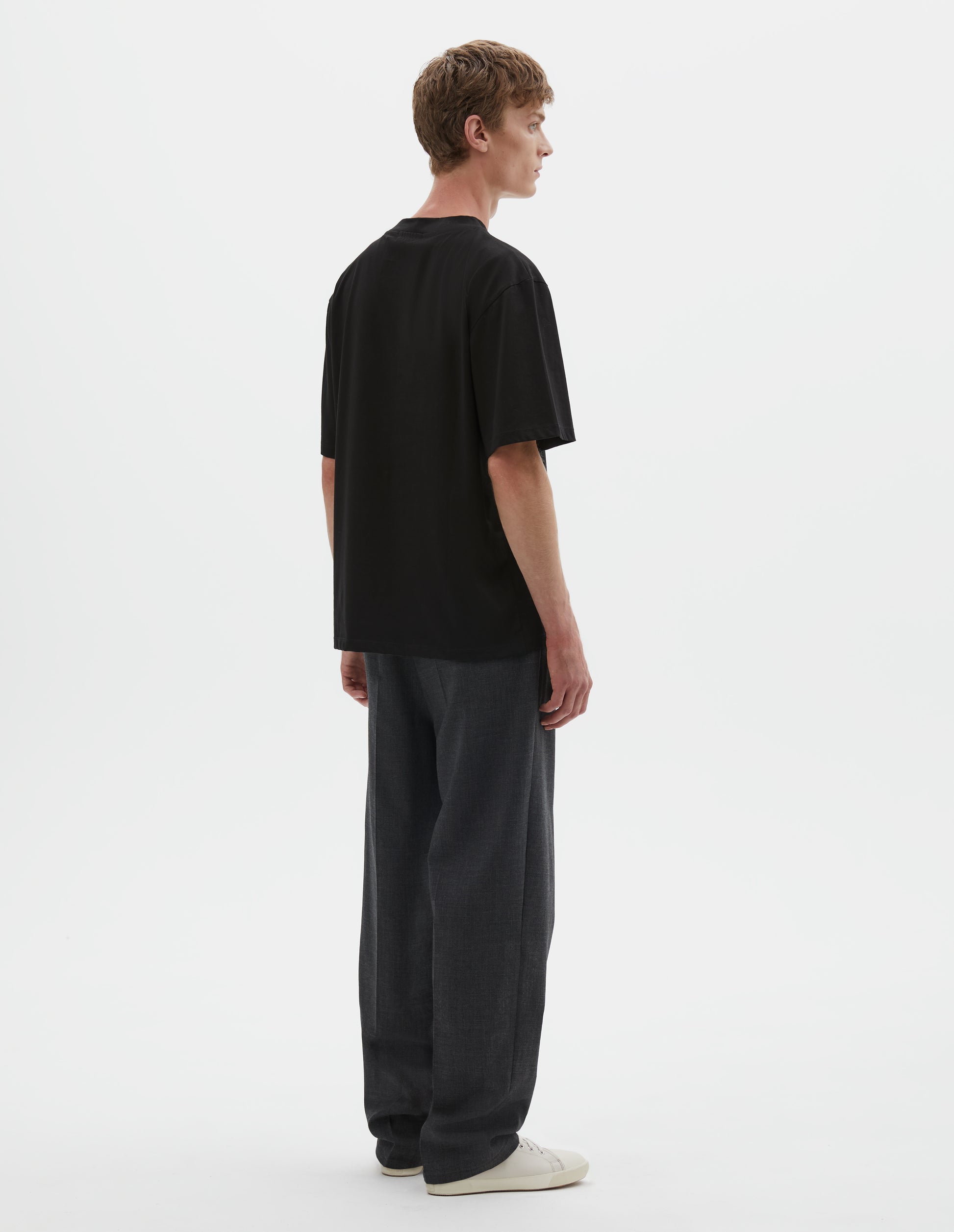 Full body image, back view of model wearing black cotton short sleeve t-shirt. Styled with grey trousers and beige canvas trainers.