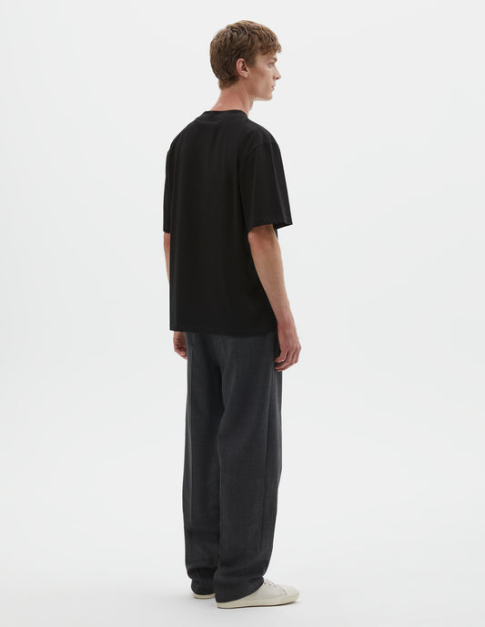 Full body image, back view of model wearing black cotton short sleeve t-shirt. Styled with grey trousers and beige canvas trainers.