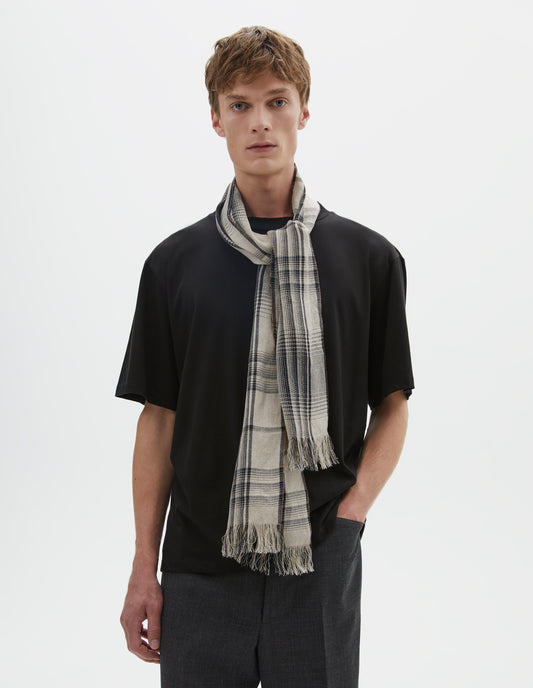 Cropped body image, front view of model wearing an off white and black checked linen scarf. Styled with a black t-shirt and dark grey trousers.