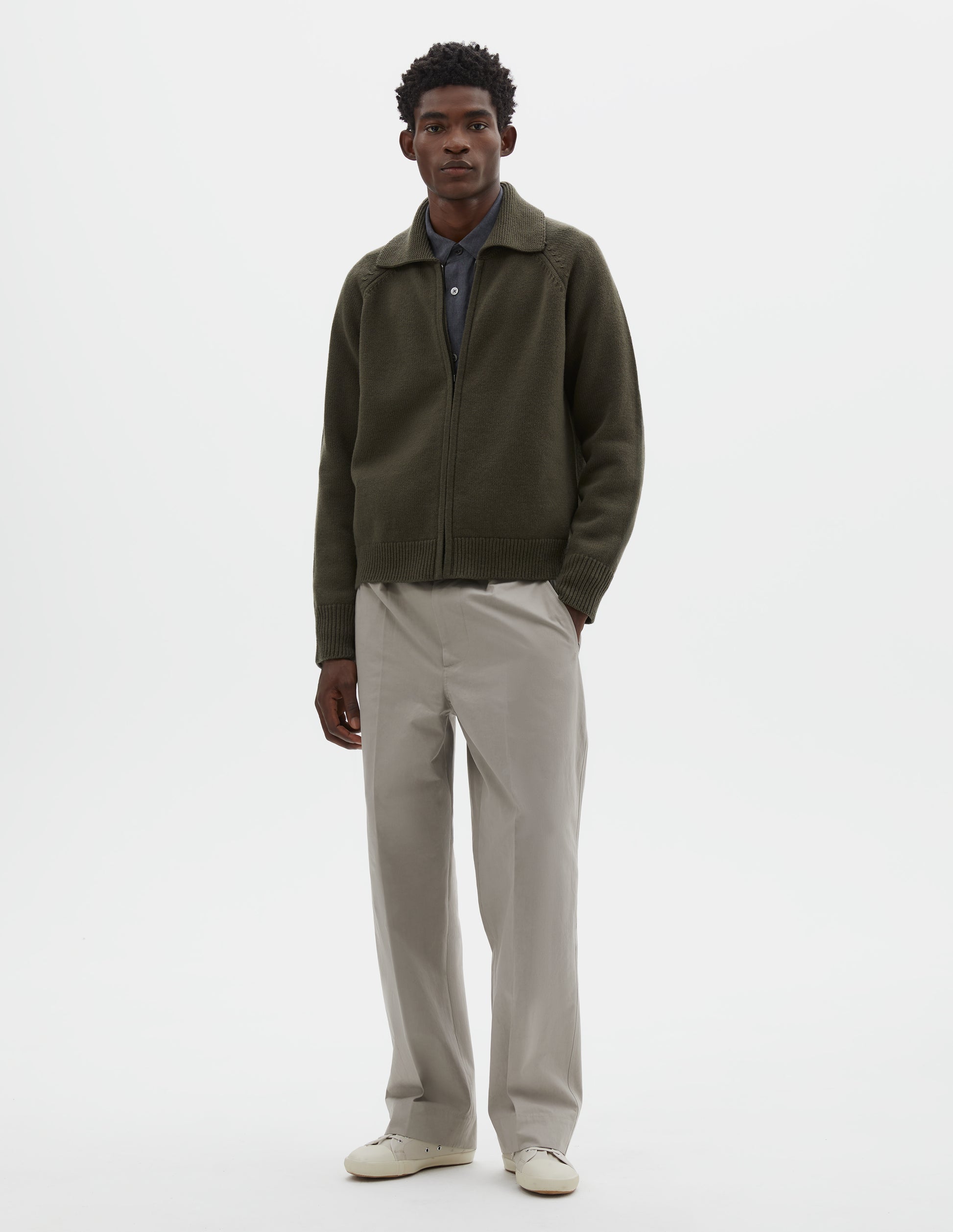 Full body image, front view of model wearing greenish brown wool cardigan, with raglan sleeve, zip fastening and contrast rib on collar, cuffs and hem. Styled over grey shirt, with beige trousers and beige canvas trainers.