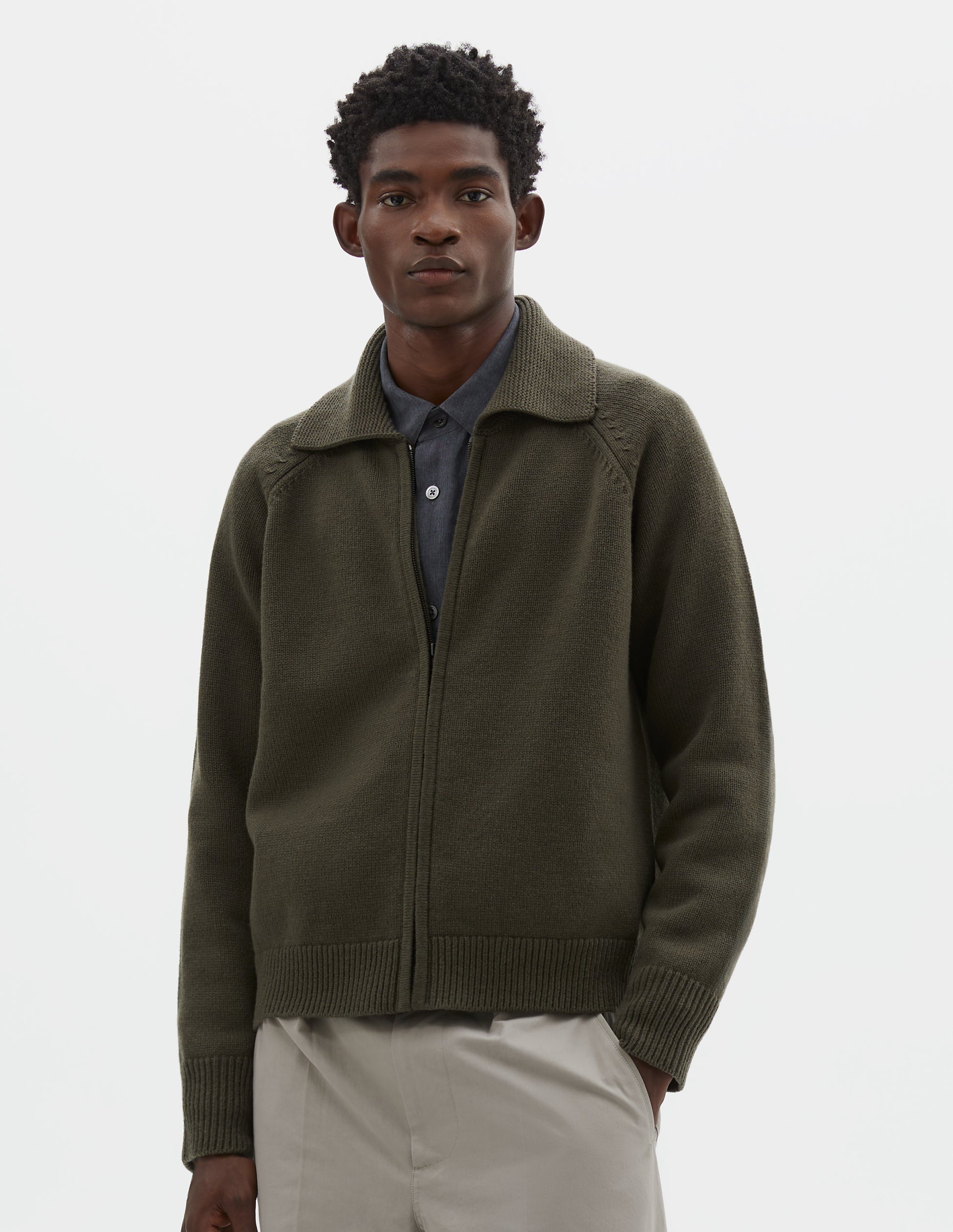 Cropped body image, front view of model wearing greenish brown wool cardigan, with raglan sleeve, zip fastening and contrast rib on collar, cuffs and hem. Styled over grey shirt, with beige trousers.