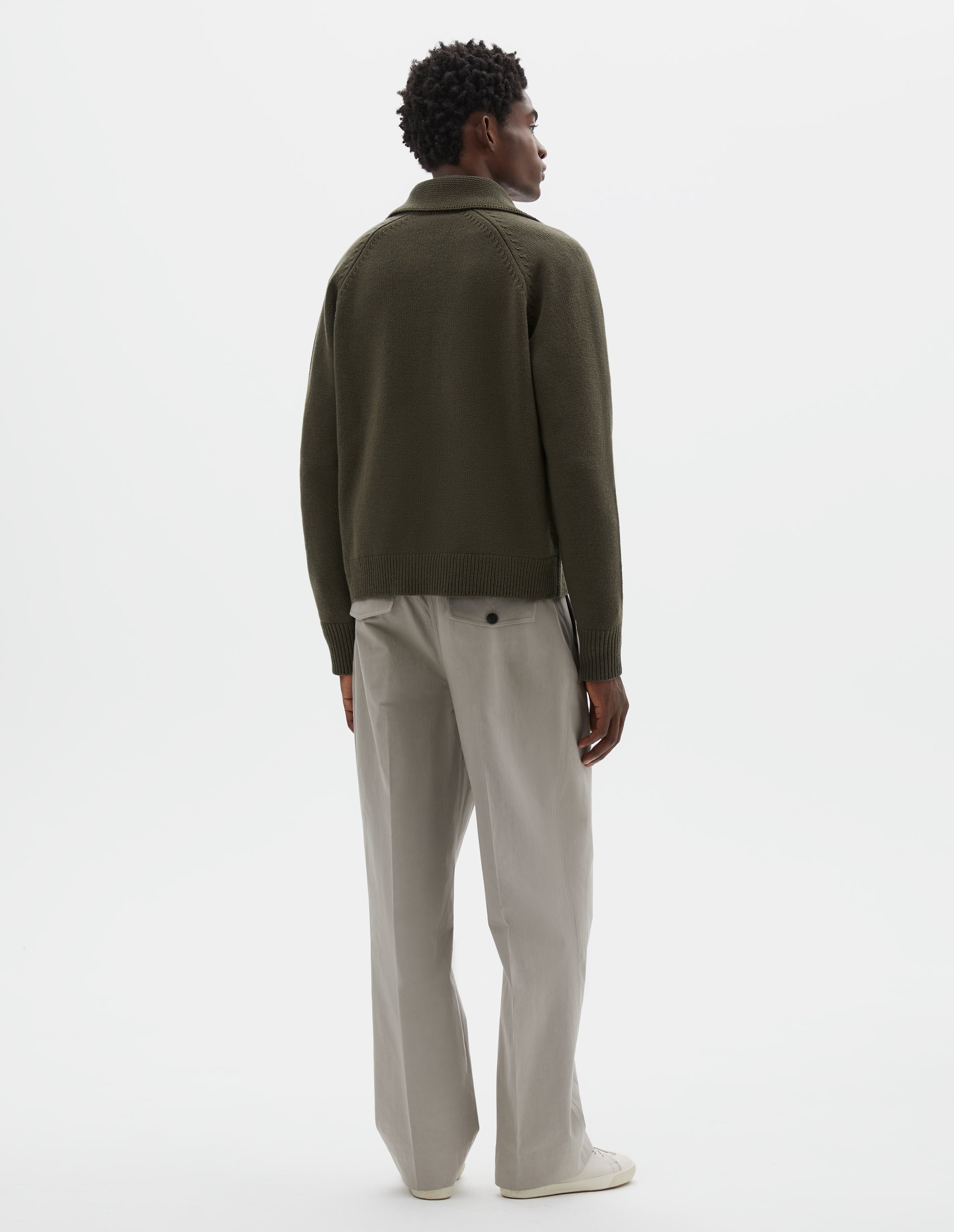 Full body image, back view of model wearing greenish brown wool cardigan, with raglan sleeve, zip fastening and contrast rib on collar, cuffs and hem. Styled with beige trousers and beige canvas trainers. 