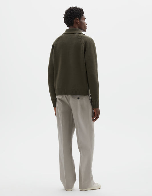 Full body image, back view of model wearing greenish brown wool cardigan, with raglan sleeve, zip fastening and contrast rib on collar, cuffs and hem. Styled with beige trousers and beige canvas trainers. 