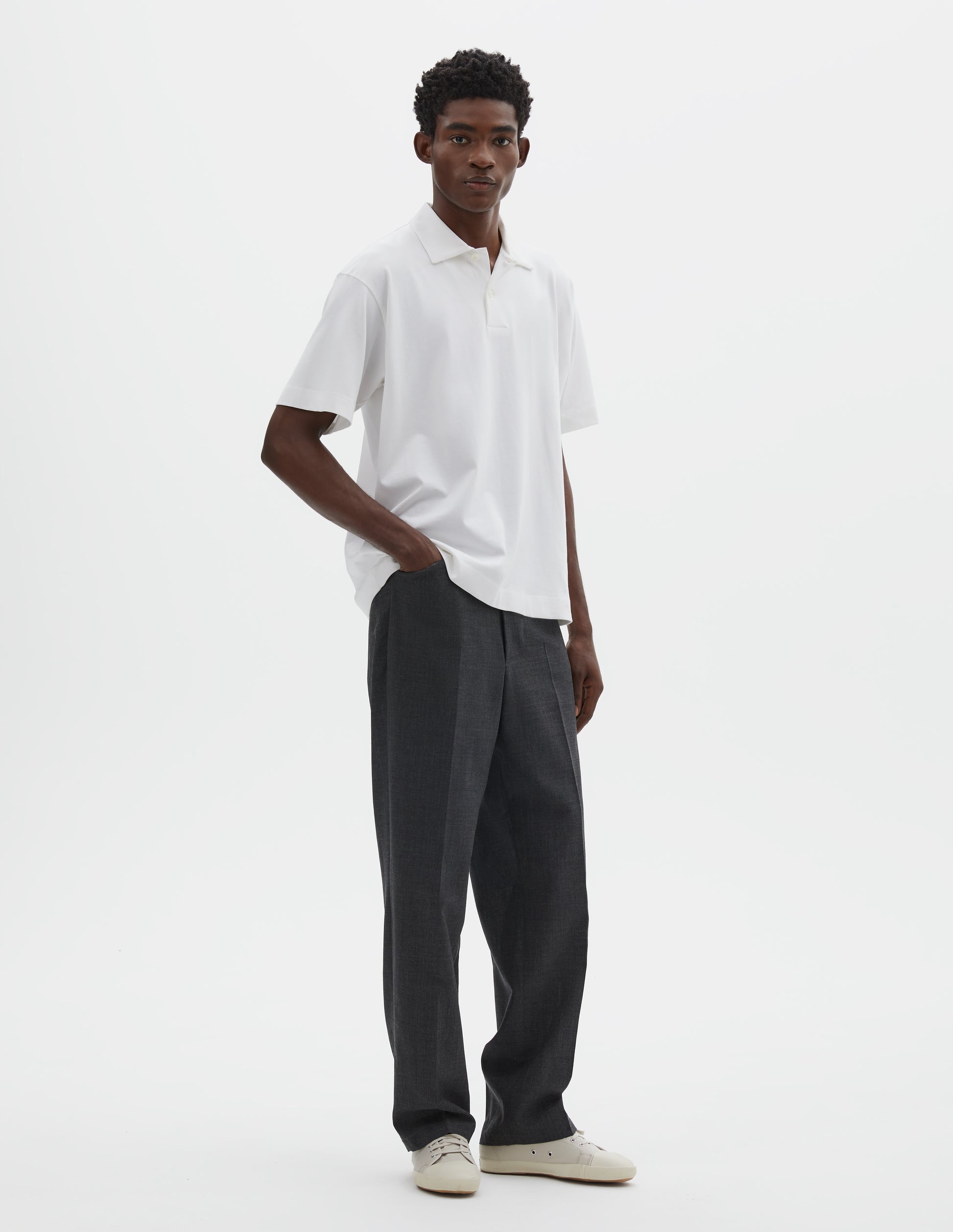 Full body image, front view of model wearing grey crisp wool trousers, with horn button fastening and slant front pockets. Styled with white short sleeve polo shirt and beige canvas trainers.