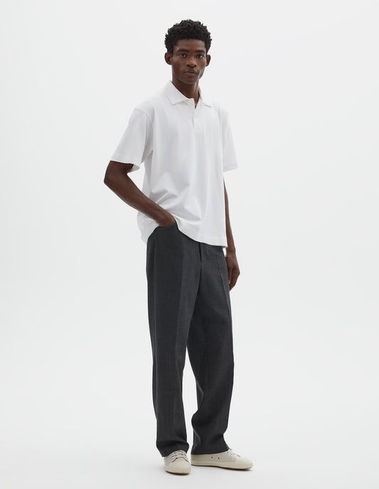 Full body image, front view of model wearing grey crisp wool trousers, with horn button fastening and slant front pockets. Styled with white short sleeve polo shirt and beige canvas trainers.