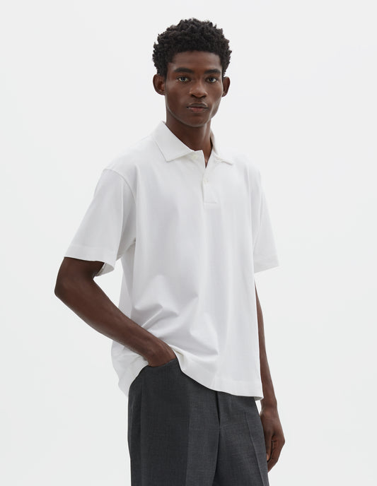 Cropped image, front view of model wearing white short sleeve cotton polo shirt with wide placket and horn buttons. Styled with dark grey trousers.