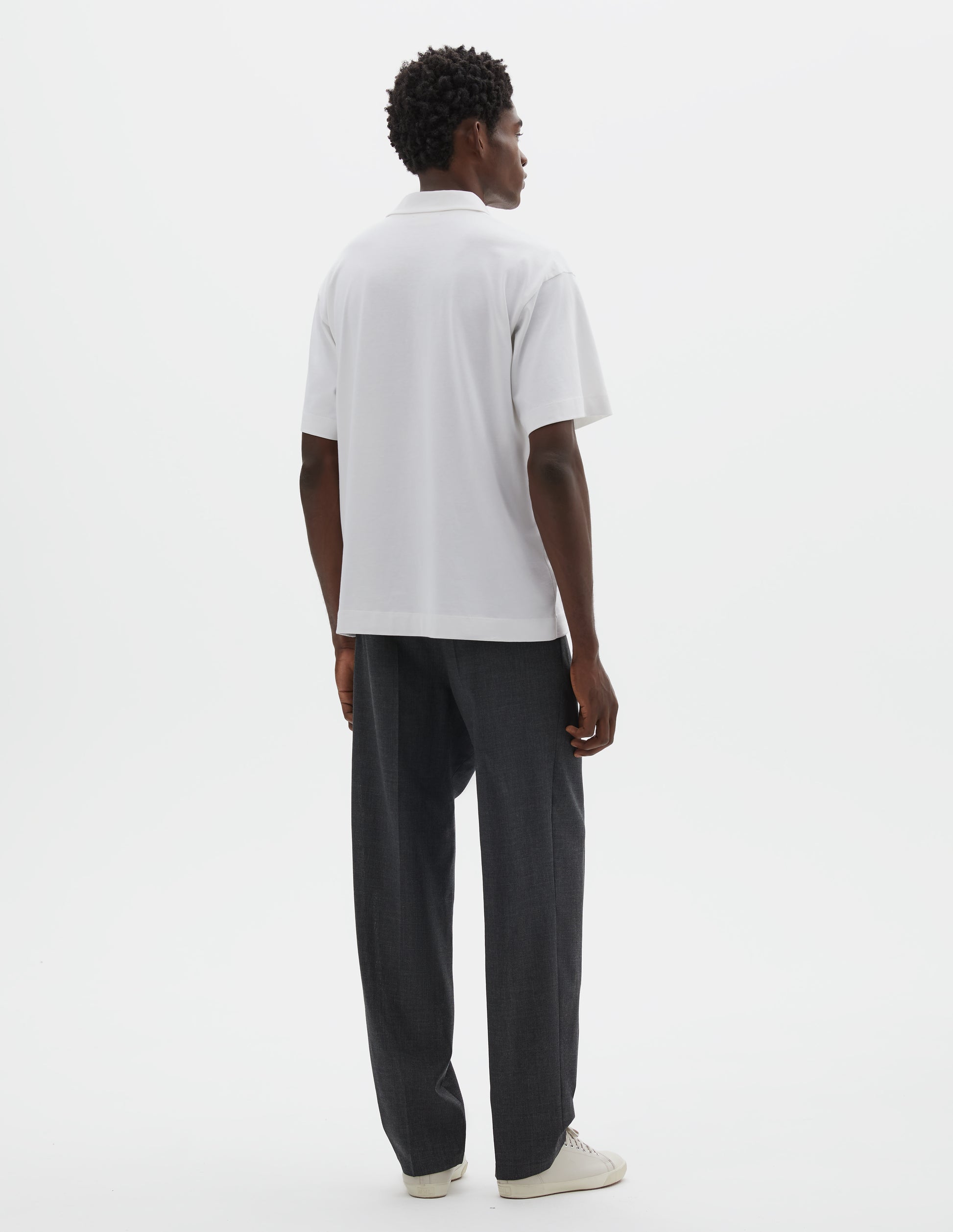 Full body image, back view of model wearing grey crisp wool trousers. Styled with white short sleeve polo shirt and beige canvas trainers.