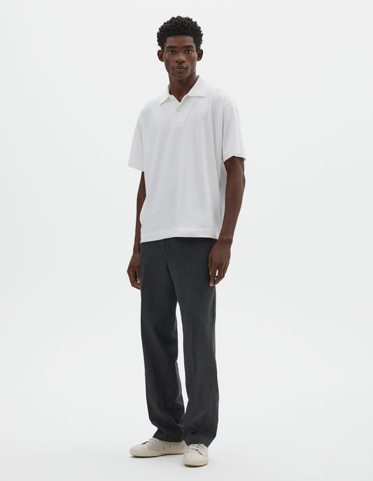 Full body image, front view of model wearing white cotton short sleeve polo shirt with wide placket and horn buttons. Styled with dark grey trousers and white canvas trainers.