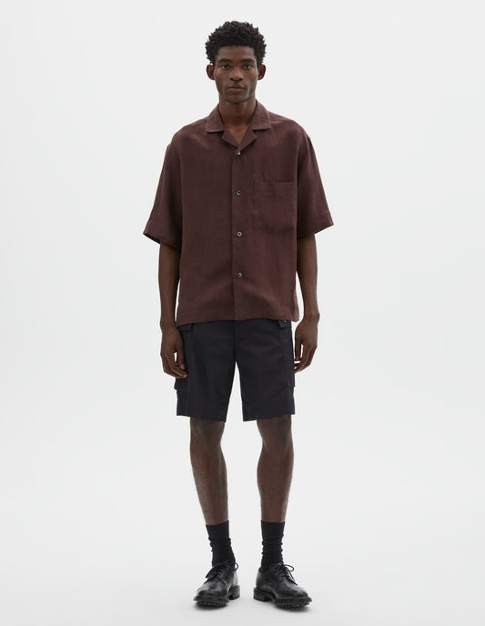 Full body image, front view of model wearing relaxed boxy short-sleeve brown shirt with revere collar, mother of pearl buttons, chest patch pocket and side split hem Styled with black cargo shorts, black socks and black leather shoes.