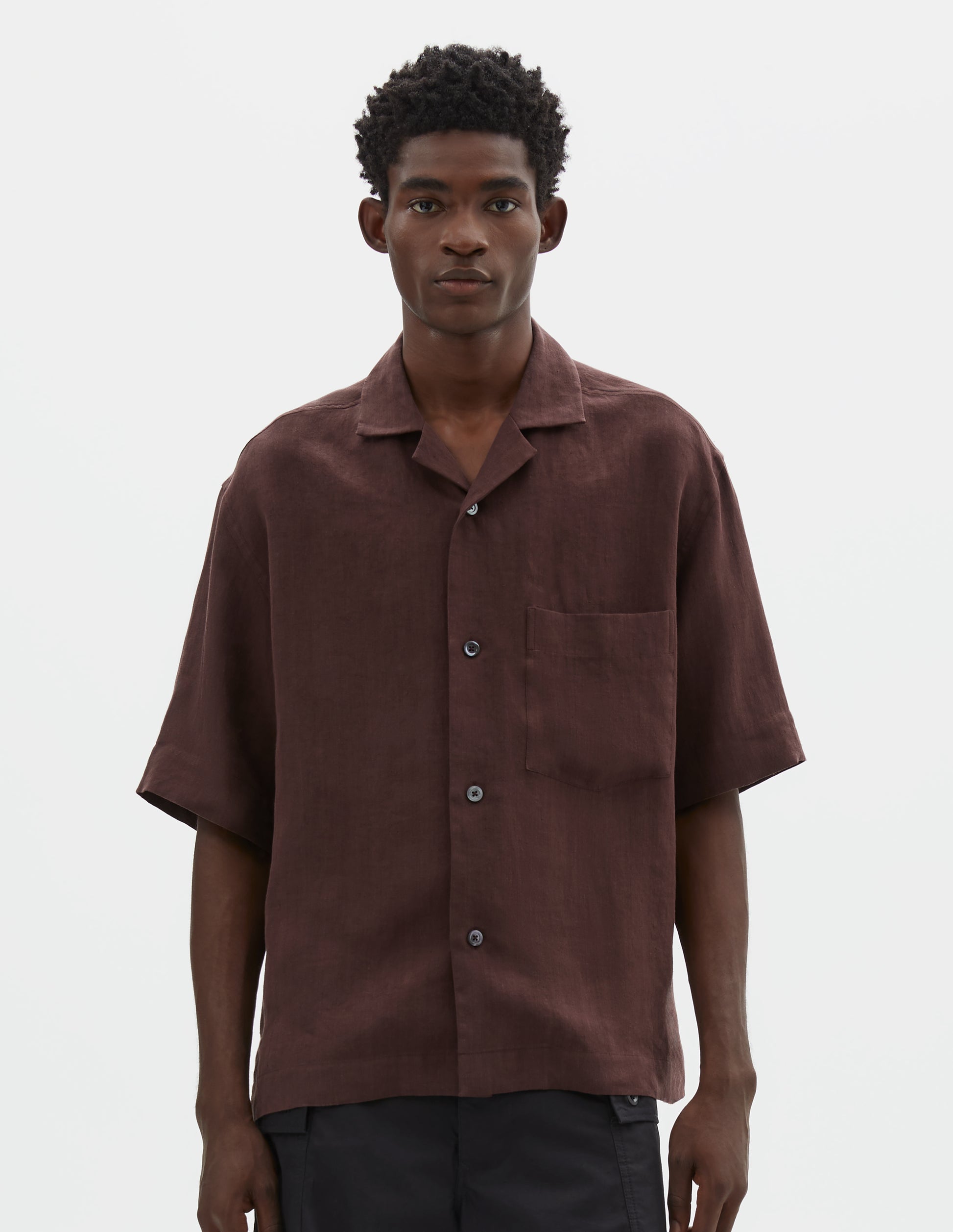 Cropped body image, front view of model wearing relaxed boxy short-sleeve brown shirt with revere collar, mother of pearl buttons, chest patch pocket and side split hem Styled with black cargo shorts.