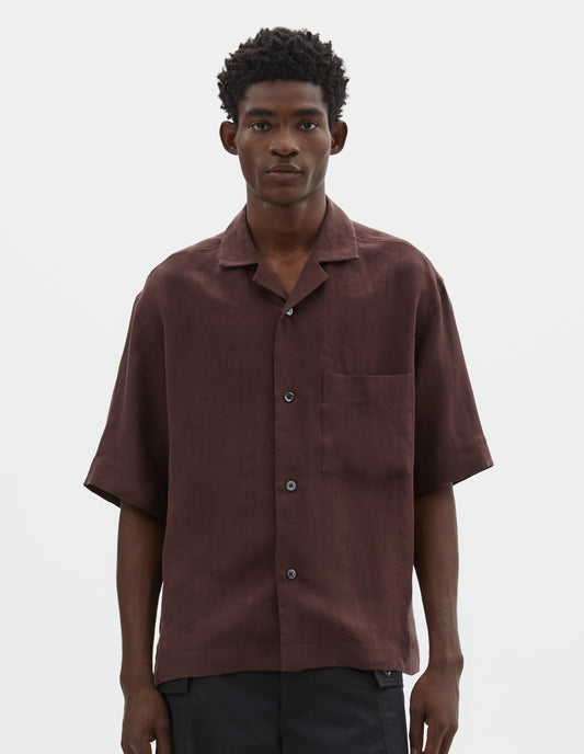 Cropped body image, front view of model wearing relaxed boxy short-sleeve brown shirt with revere collar, mother of pearl buttons, chest patch pocket and side split hem Styled with black cargo shorts.