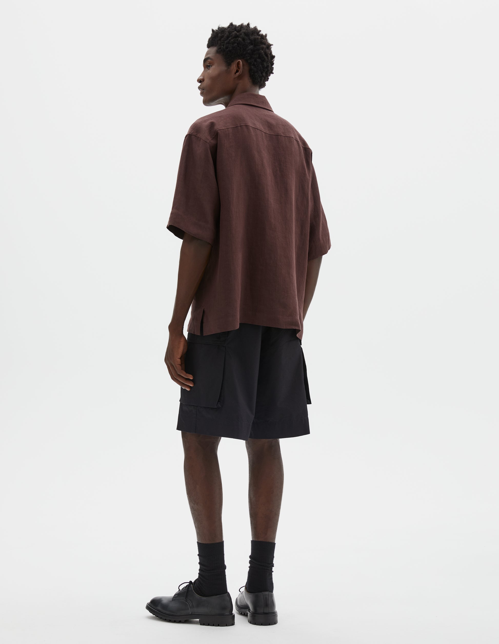 Full body image, back view of model wearing relaxed boxy short-sleeve brown shirt with side split hem. Styled with black cargo shorts, black socks and black leather shoes.