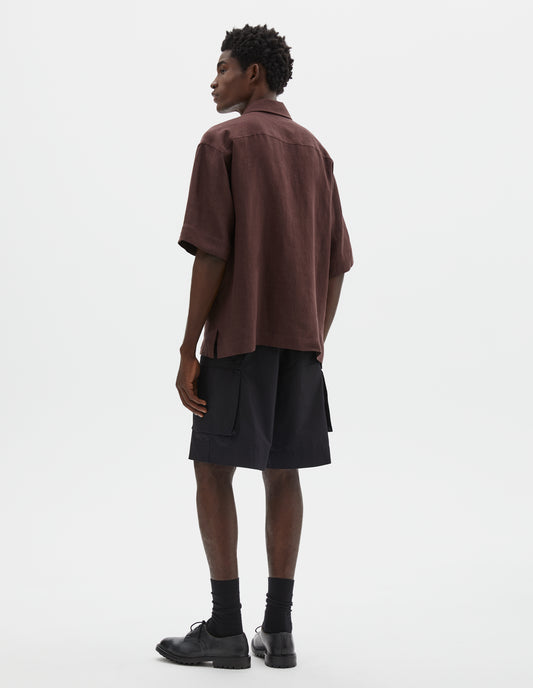 Full body image, back view of model wearing relaxed boxy short-sleeve brown shirt with side split hem. Styled with black cargo shorts, black socks and black leather shoes.