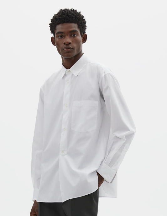 Cropped body image, front view of model wearing white cotton shirt with chest pocket, curved hem, curved placket from waist to hem and mother of pearl buttons. Styled with dark grey pleated trousers.