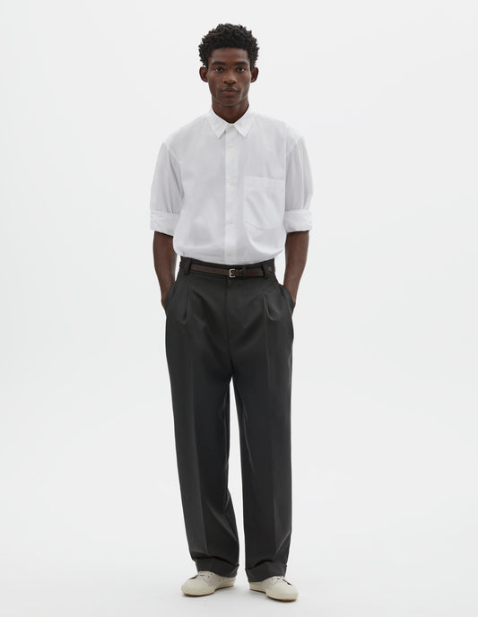 Full body image, front view of model wearing dark grey trousers, relaxed double pleat with horn button waistband, belt loops and cuffed hem. Styled with white shirt, dark brown belt and beige trainers.