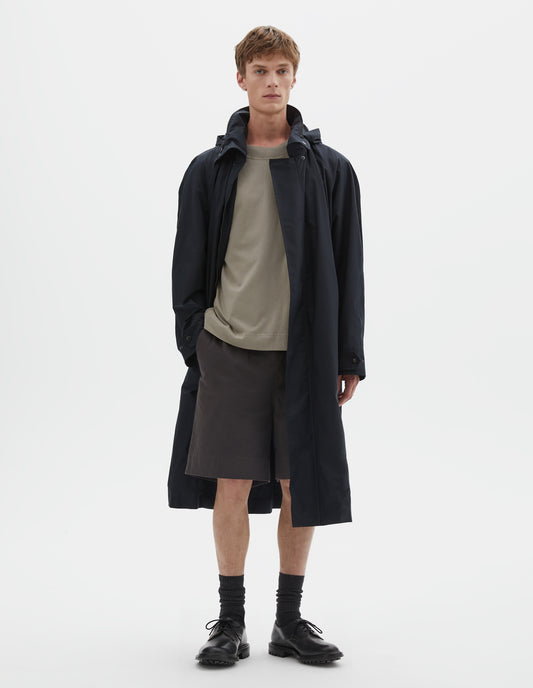 Full body image, front view of model wearing black ventile cotton poplin longline mac with slant pockets, vislon zip and adjustable popper cuffs. Styled with beige t-shirt, grey shorts, black socks and black leather brogues.