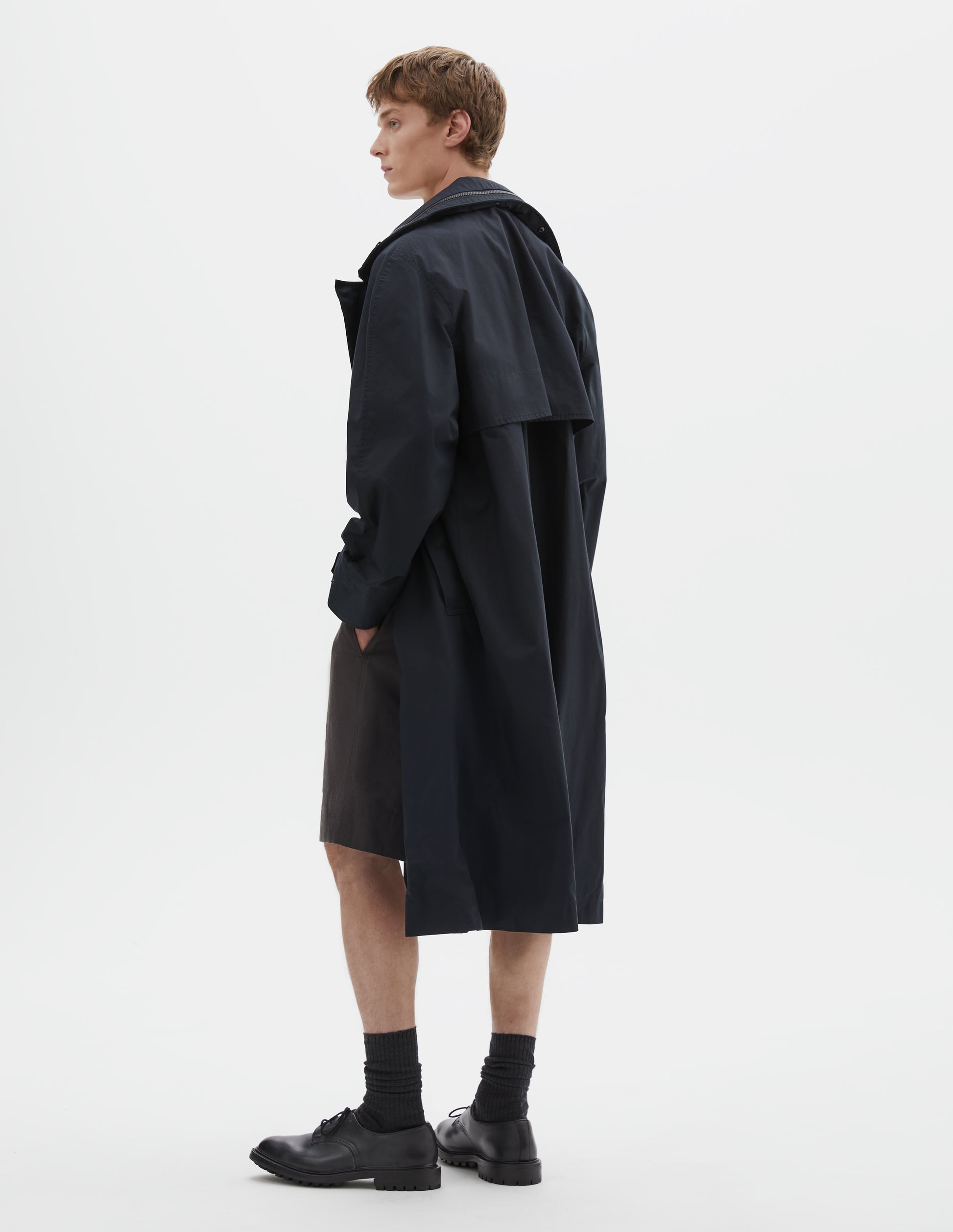 Full body image, back view of model wearing black ventile cotton poplin longline mac with deep storm vent and packable hood. Styled with grey shorts, black socks and black leather brogues.