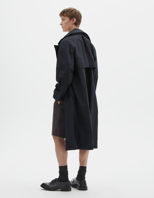 Full body image, back view of model wearing black ventile cotton poplin longline mac with deep storm vent and packable hood. Styled with grey shorts, black socks and black leather brogues.