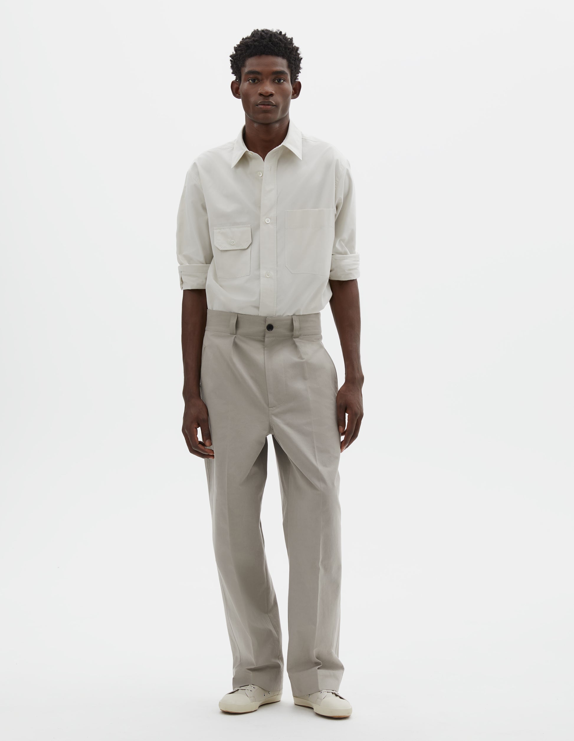 Full body image, front view of model wearing light grey cotton trousers, with single pleats, horn button closure, slant jet pockets and belt loops. Styled with white shirt tucked and off white canvas trainers.