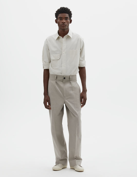 Full body image, front view of model wearing light grey cotton trousers, with single pleats, horn button closure, slant jet pockets and belt loops. Styled with white shirt tucked and off white canvas trainers.