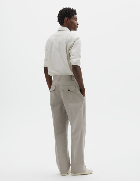 Full body image, back view of model wearing light grey cotton trousers, with belt loops and concealed flap and button-through jet back pockets. Styled with white shirt tucked and off white canvas trainers.