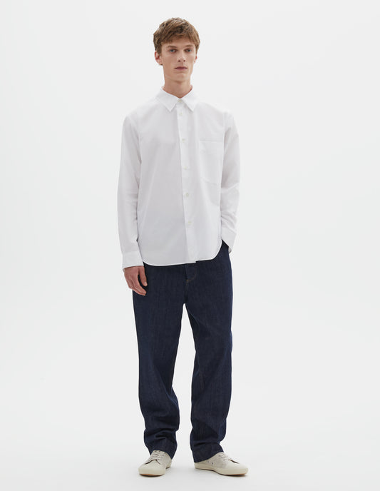 Full body image, front view of model wearing white cotton shirt with chest pocket, curved hem and mother of pearl buttons. Styled with indigo blue jeans and beige trainers.
