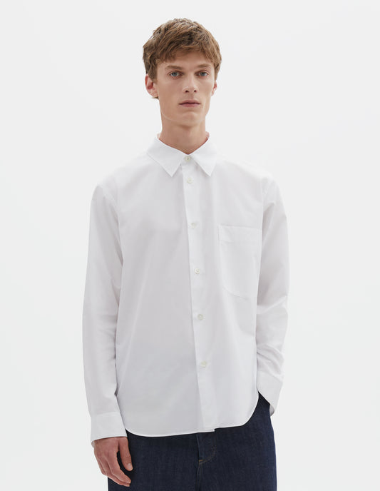 Cropped body image, front view of model wearing white cotton shirt with chest pocket, curved hem and mother of pearl buttons. Styled with indigo blue jeans.