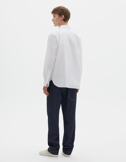 Full body image, back view of model wearing white cotton shirt, with curved hem, pleats on back yoke and mother of pearl button cuffs. Styled with indigo blue jeans and beige trainers.