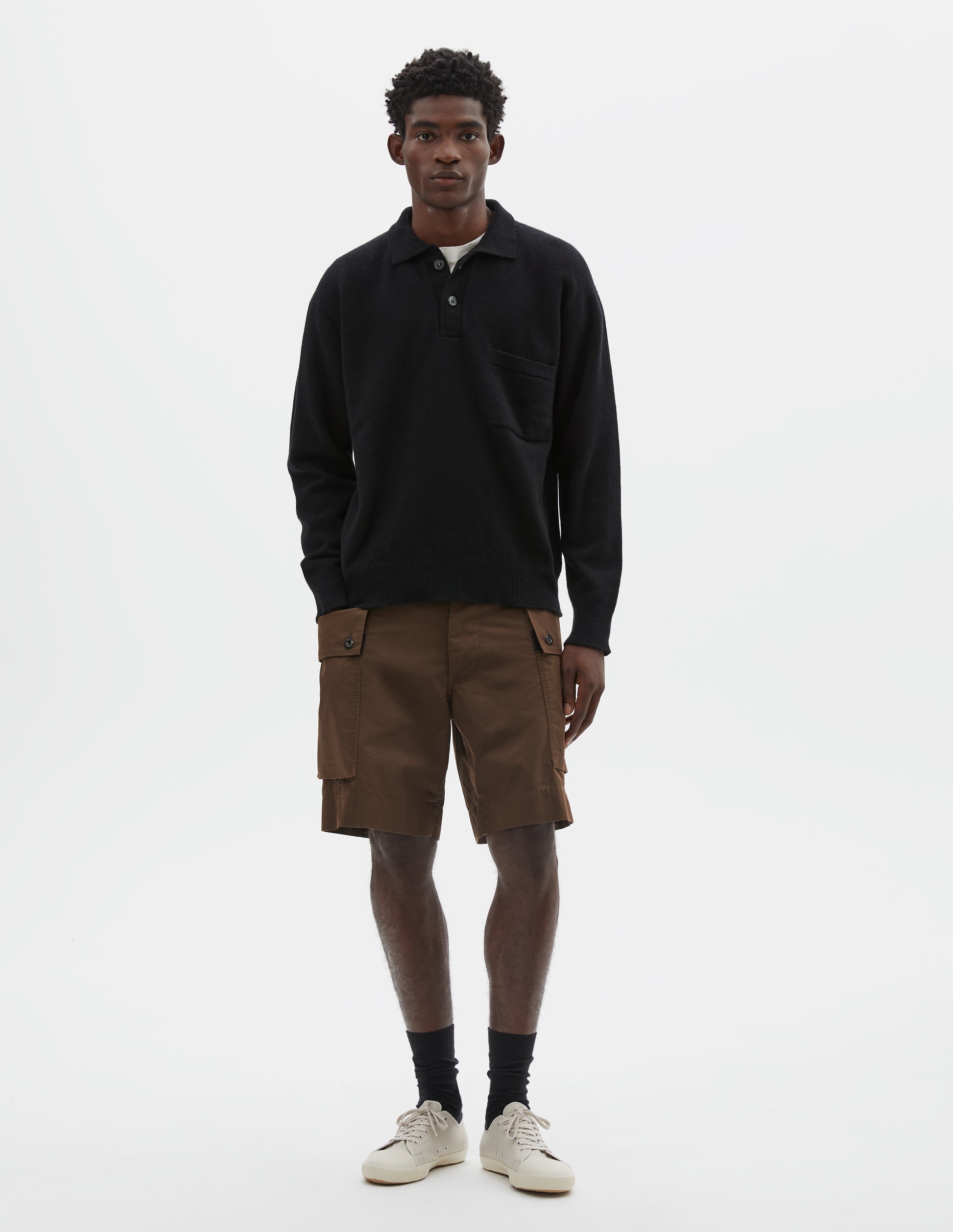 Full body image, front view of model wearing black merino cashmere polo shirt with dropped sleeve, integral chest pocket, horn button closure and rib collar, cuffs and hem. Styled over white t-shirt, with brown cargo shorts, black socks and beige canvas trainers.
