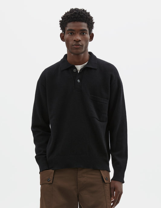 Cropped body image, front view of model wearing black merino cashmere polo shirt with dropped sleeve, integral chest pocket, horn button closure and rib collar, cuffs and hem. Styled over white t-shirt, with brown cargo shorts.