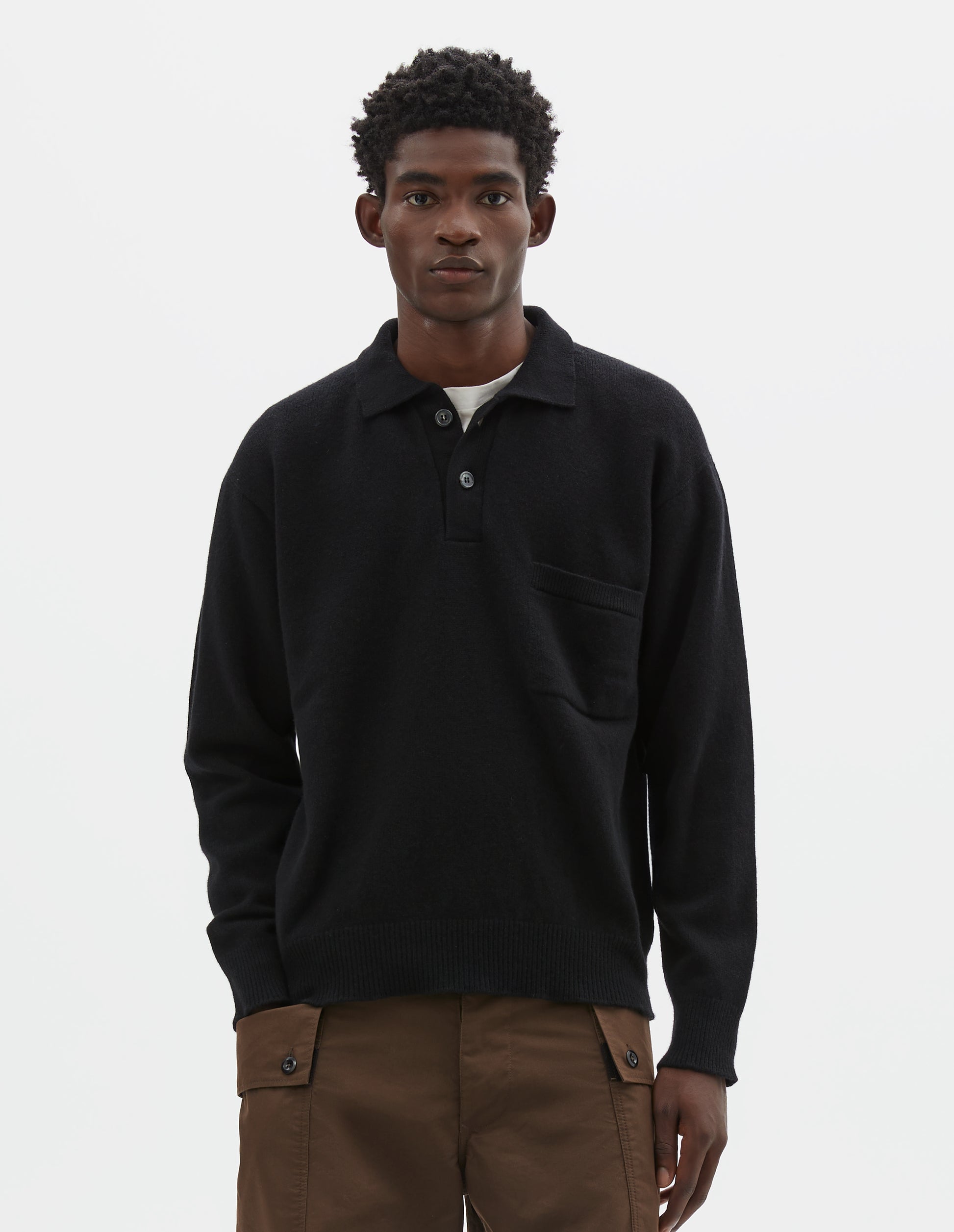 Cropped body image, front view of model wearing black merino cashmere polo shirt with dropped sleeve, integral chest pocket, horn button closure and rib collar, cuffs and hem. Styled over white t-shirt, with brown cargo shorts.