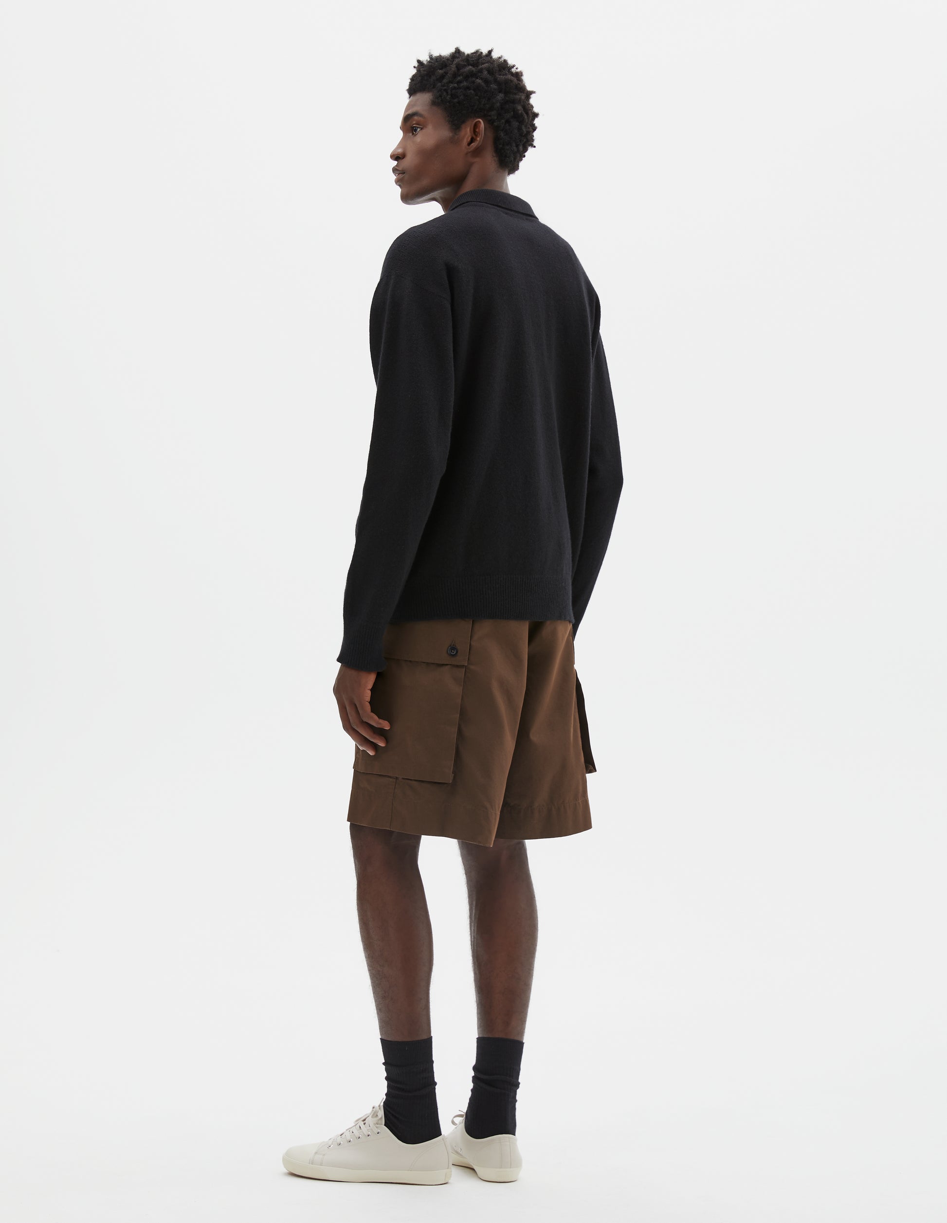 Full body image, back view of model wearing black merino cashmere polo shirt with dropped sleeve and rib collar, cuffs and hem. Styled with brown cargo shorts, black socks and beige canvas trainers.