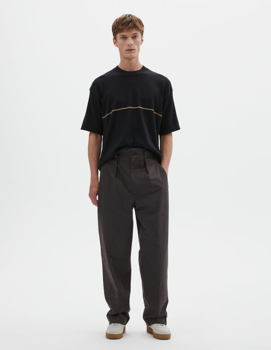 Full body image, front view of model wearing dark brown cotton trousers with single inverted pleat, deep stitch hem, slant jet pockets and belt loops. Styled with black t shirt with yellow line across chest and white trainers.