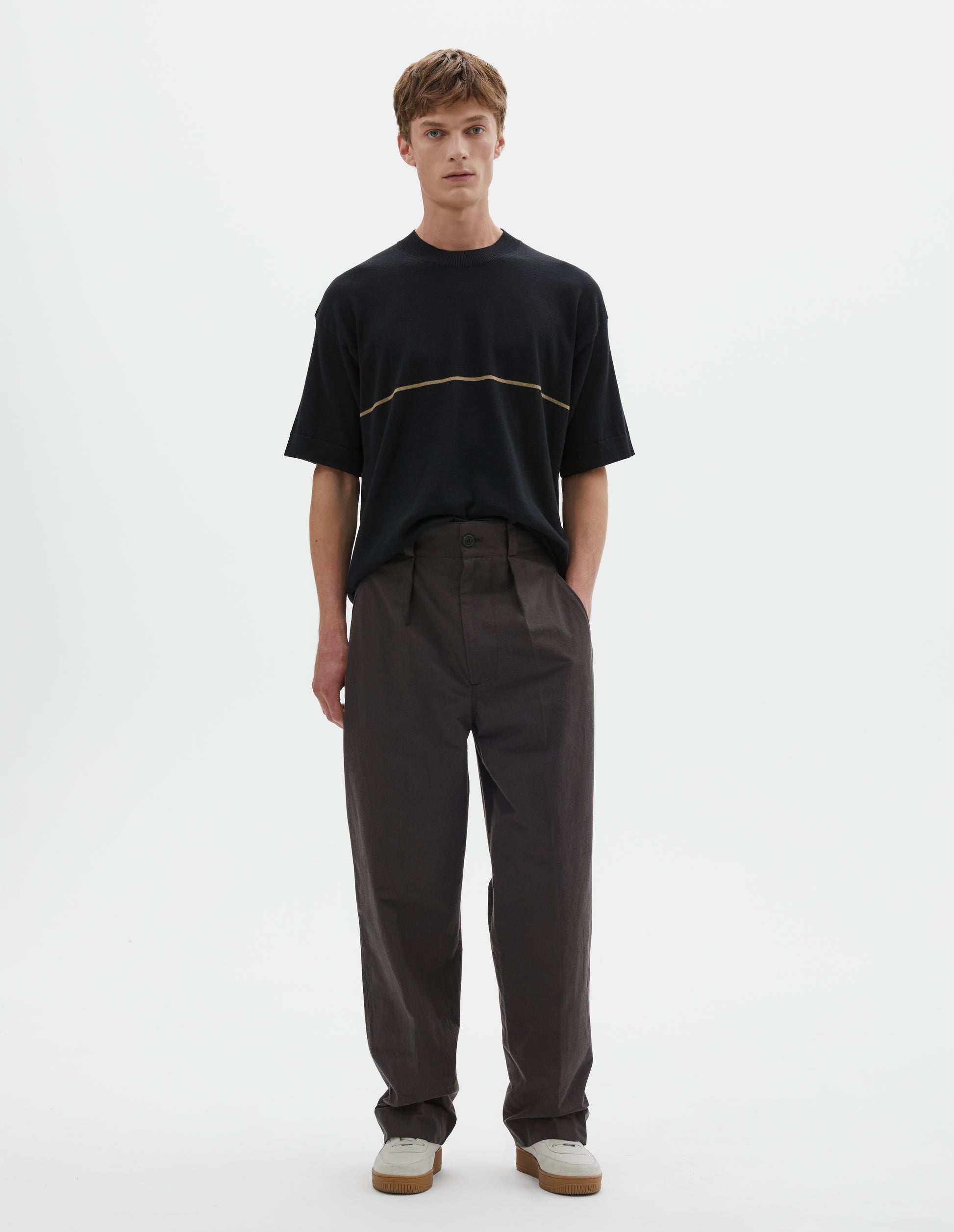 Full body image, front view of model wearing dark brown cotton trousers with single inverted pleat, deep stitch hem, slant jet pockets and belt loops. Styled with black t shirt with yellow line across chest and white trainers. 
