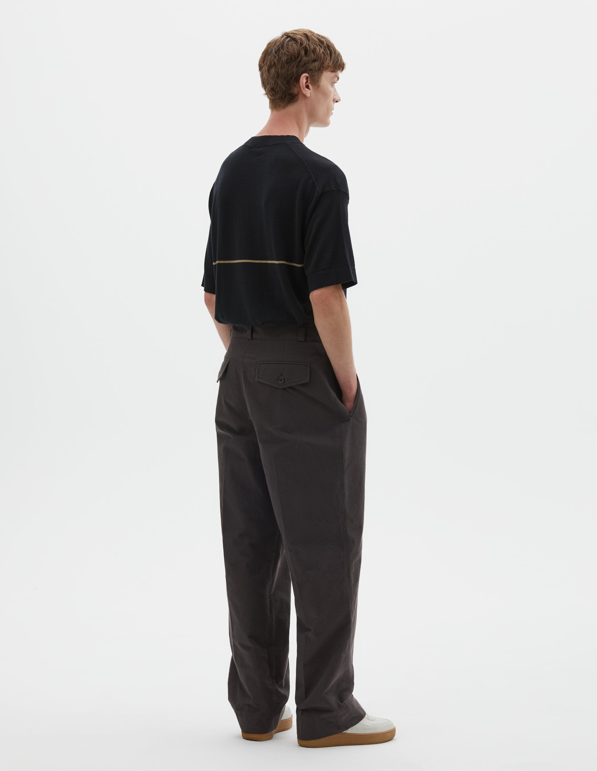 Full body image, back view of model wearing dark brown cotton trousers with belt loops and concealed and button through jet back pockets. Styled with black t shirt with yellow line across back and white trainers. 