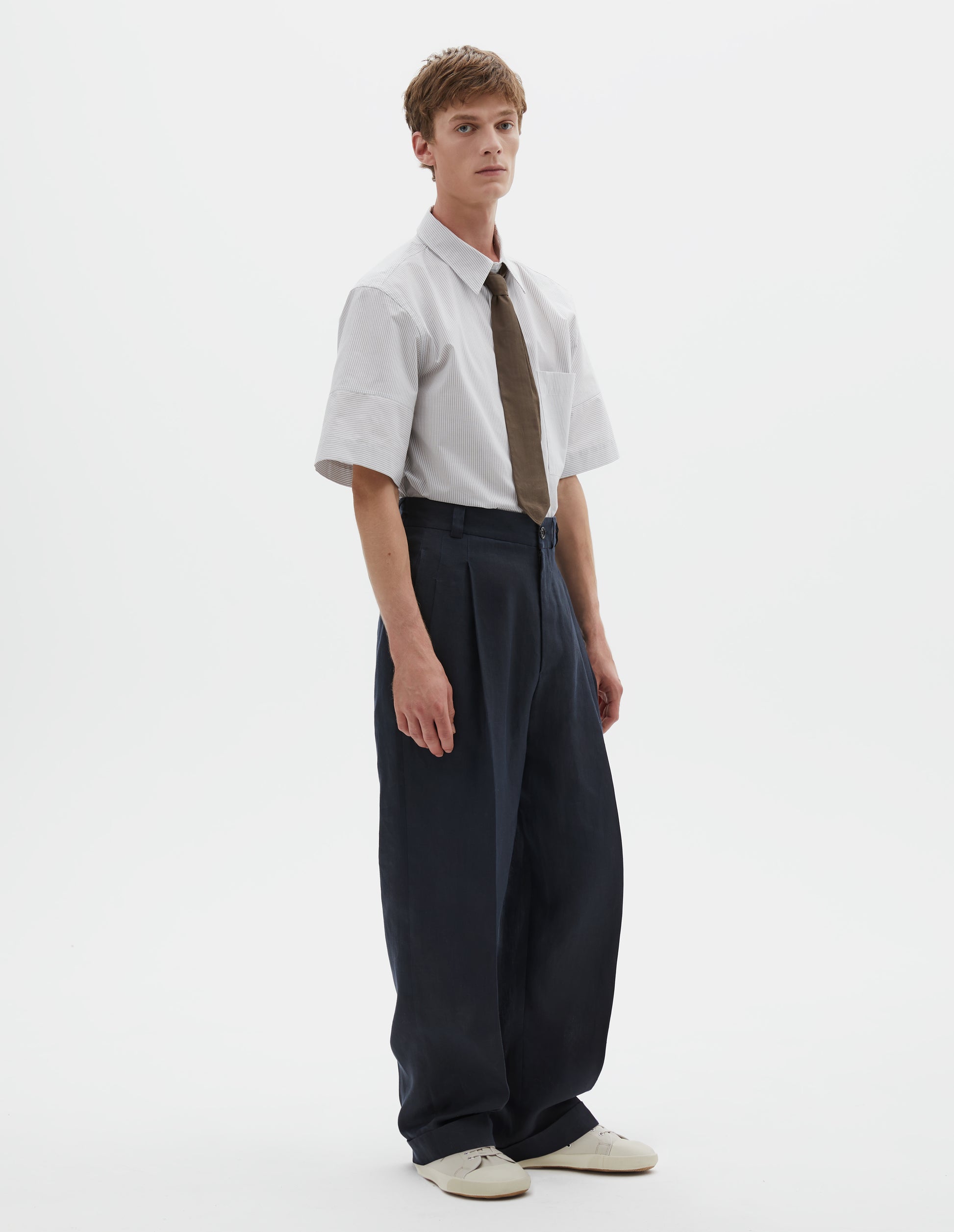 Full body image, front view of model wearing dark navy trousers, relaxed double pleat with horn button waistband, belt loops and cuffed hem. Styled with a white and grey short sleeve shirt tucked in and greenish brown tie, with beige canvas trainers.
