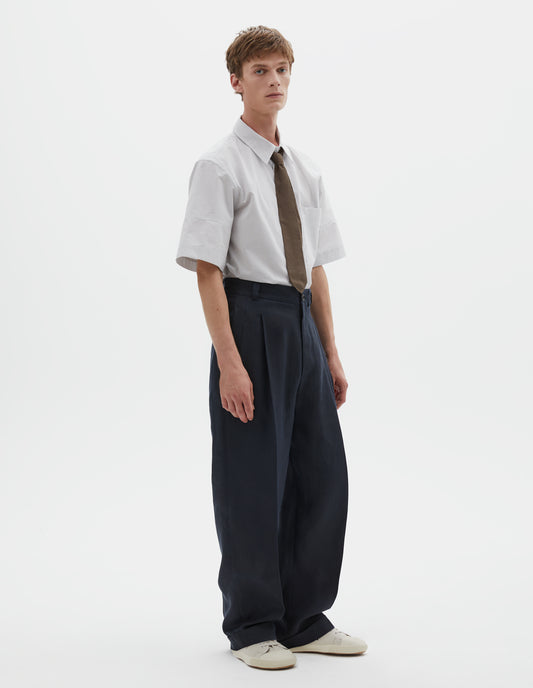 Full body image, front view of model wearing dark navy trousers, relaxed double pleat with horn button waistband, belt loops and cuffed hem. Styled with a white and grey short sleeve shirt tucked in and greenish brown tie, with beige canvas trainers.
