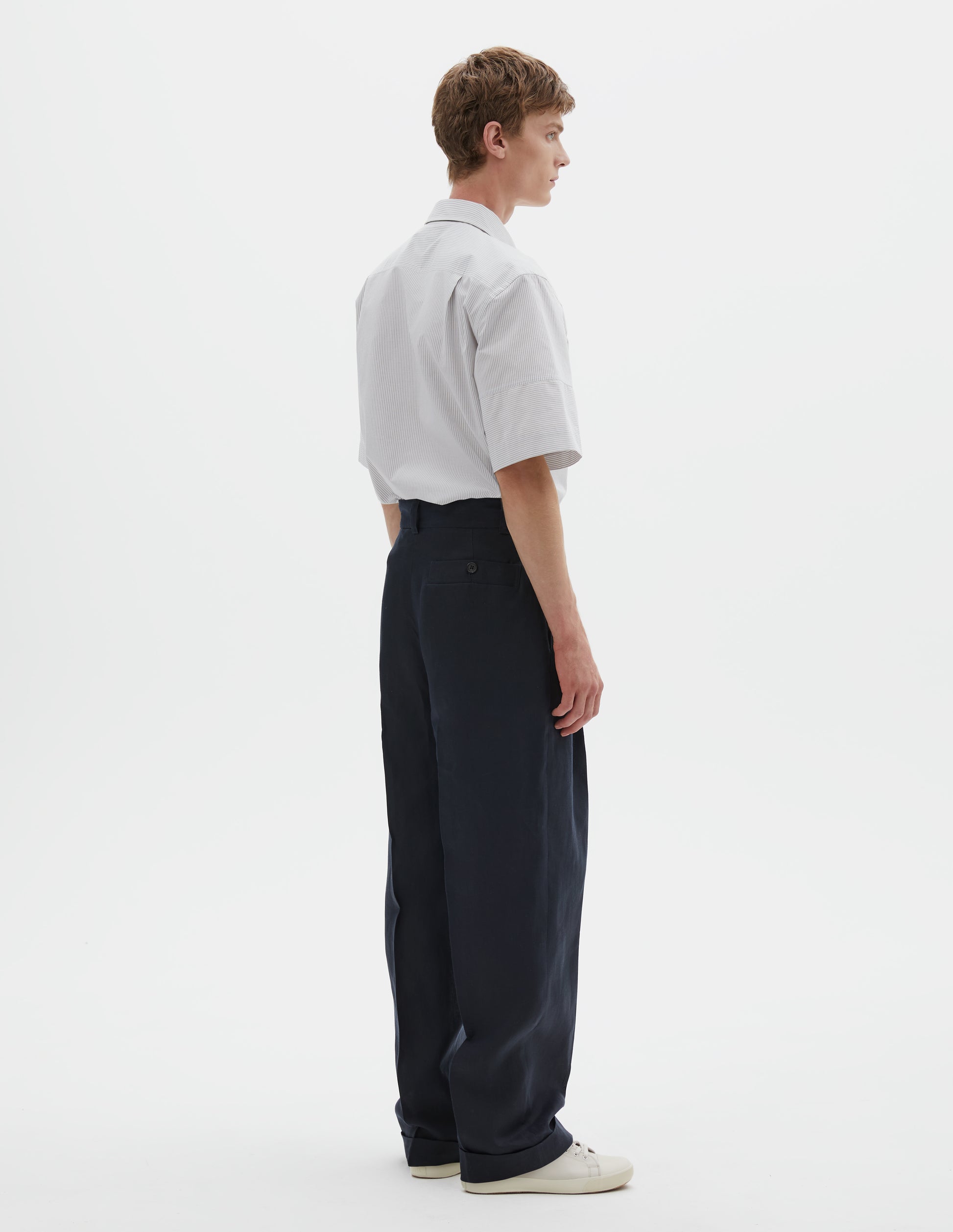 Full body image, back view of model wearing dark navy trousers, deep button-through welt back pocket, belt loops. Styled with white and grey striped short sleeve shirt tucked in and beige canvas trainers.