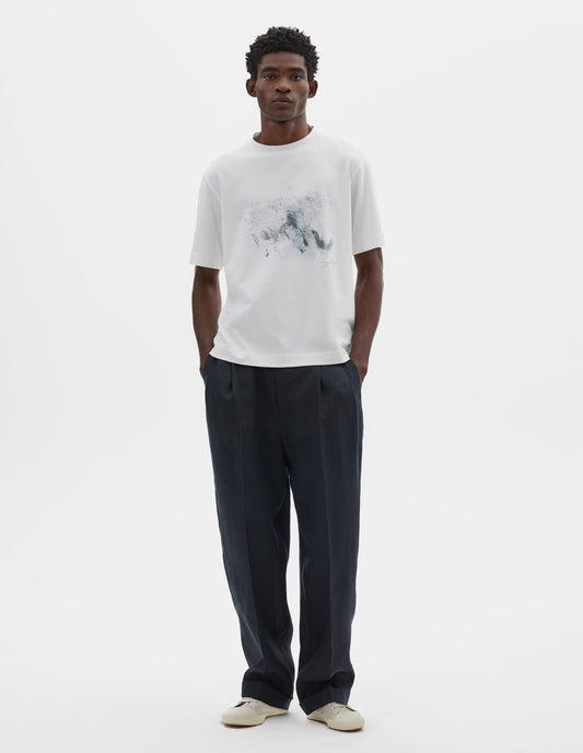 Full body image, front view of model wearing white relaxed, boxy crew neck t-shirt with dark grey drawing print. Styled with dark blue  trousers and beige canvas trainers.