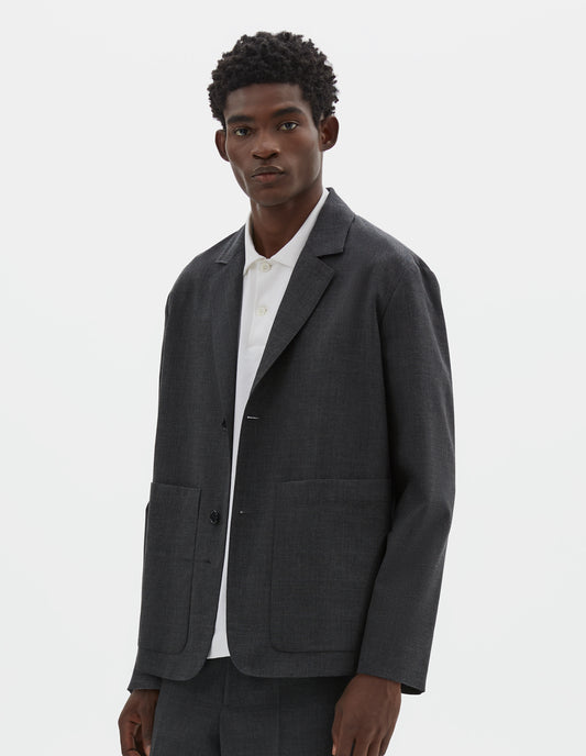 Cropped body image, front view of model wearing grey crisp wool blazer with horn button fastening and patch pockets. Styled over white shirt, with matching grey trousers.