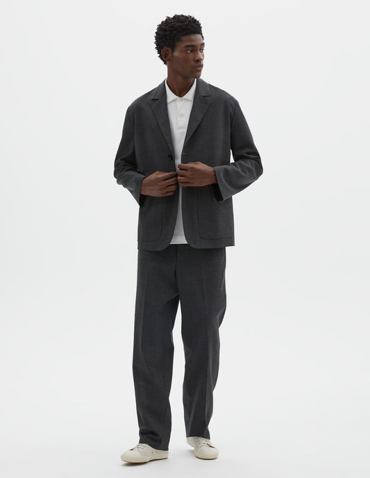 Full body image, front view of model wearing grey crisp wool blazer with horn button fastening and patch pockets. Styled over white shirt, with matching grey trousers and beige canvas trainers. 