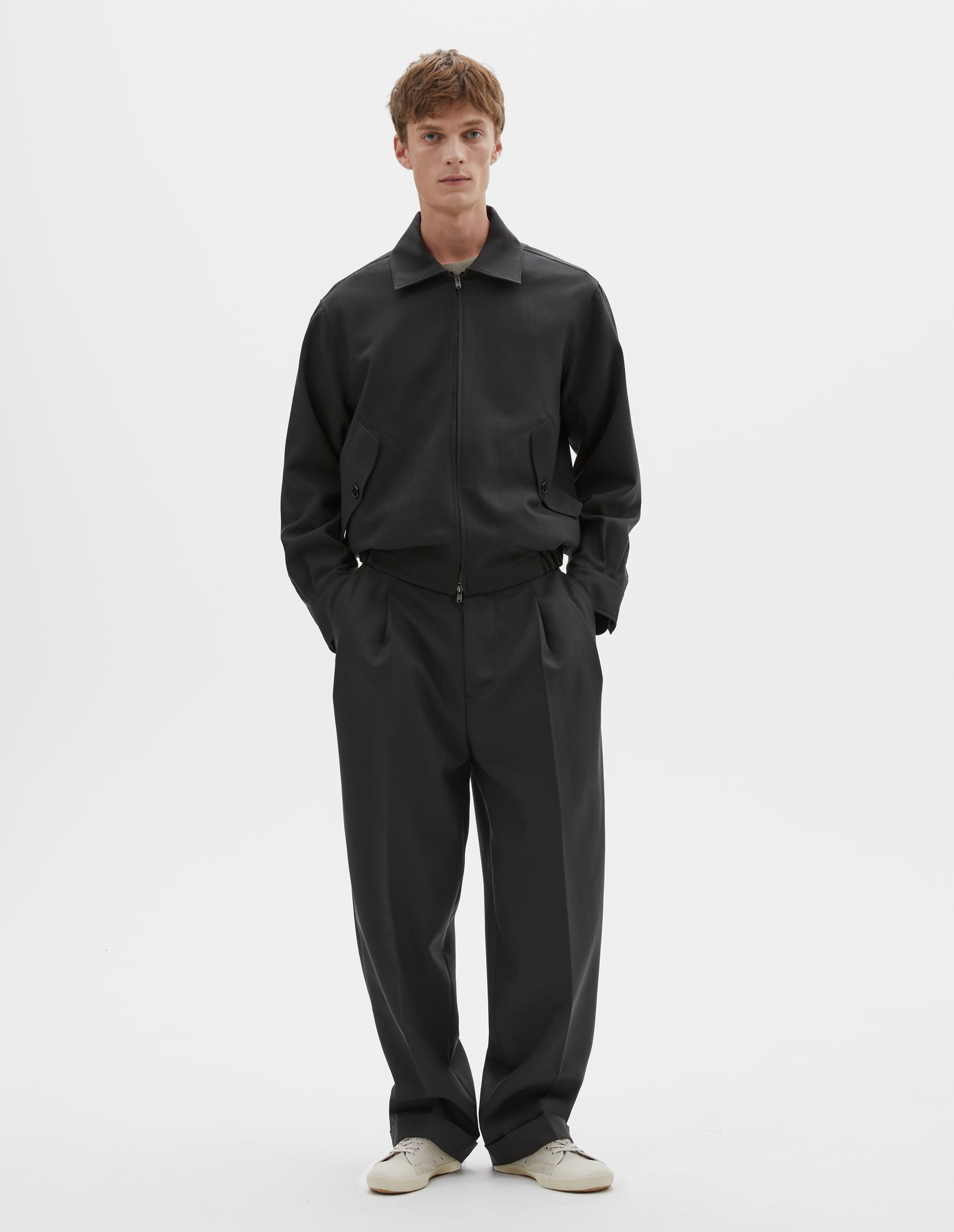 Full body image, front view of model wearing dark grey wool jacket with two way zip fastening, elasticated waistband, tab shirt cuffs and button flap pockets. Styled with black pleated trousers and white canvas trainers.