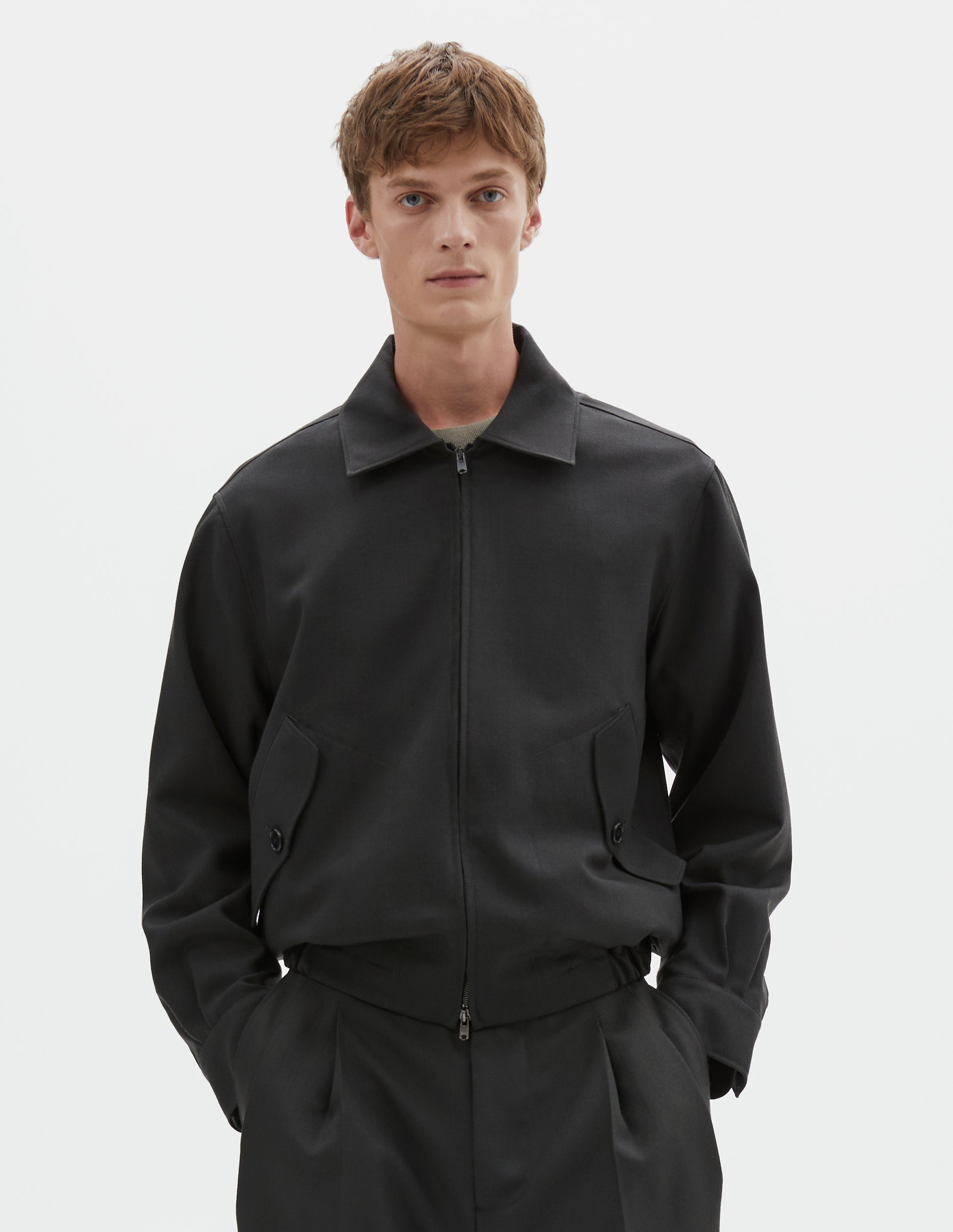 Cropped body image, front view of model wearing dark grey wool jacket with two way zip fastening, elasticated waistband, tab shirt cuffs and button flap pockets. Styled with black pleated trousers.