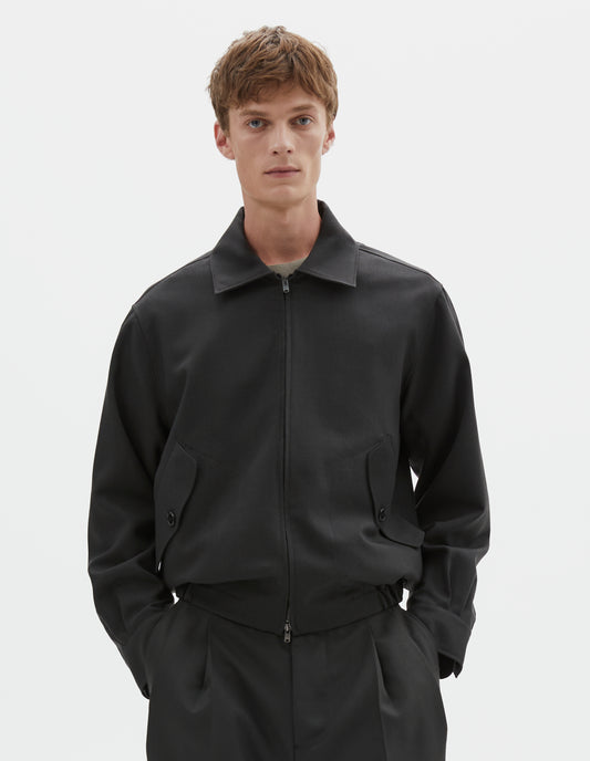 Cropped body image, front view of model wearing dark grey wool jacket with two way zip fastening, elasticated waistband, tab shirt cuffs and button flap pockets. Styled with black pleated trousers.