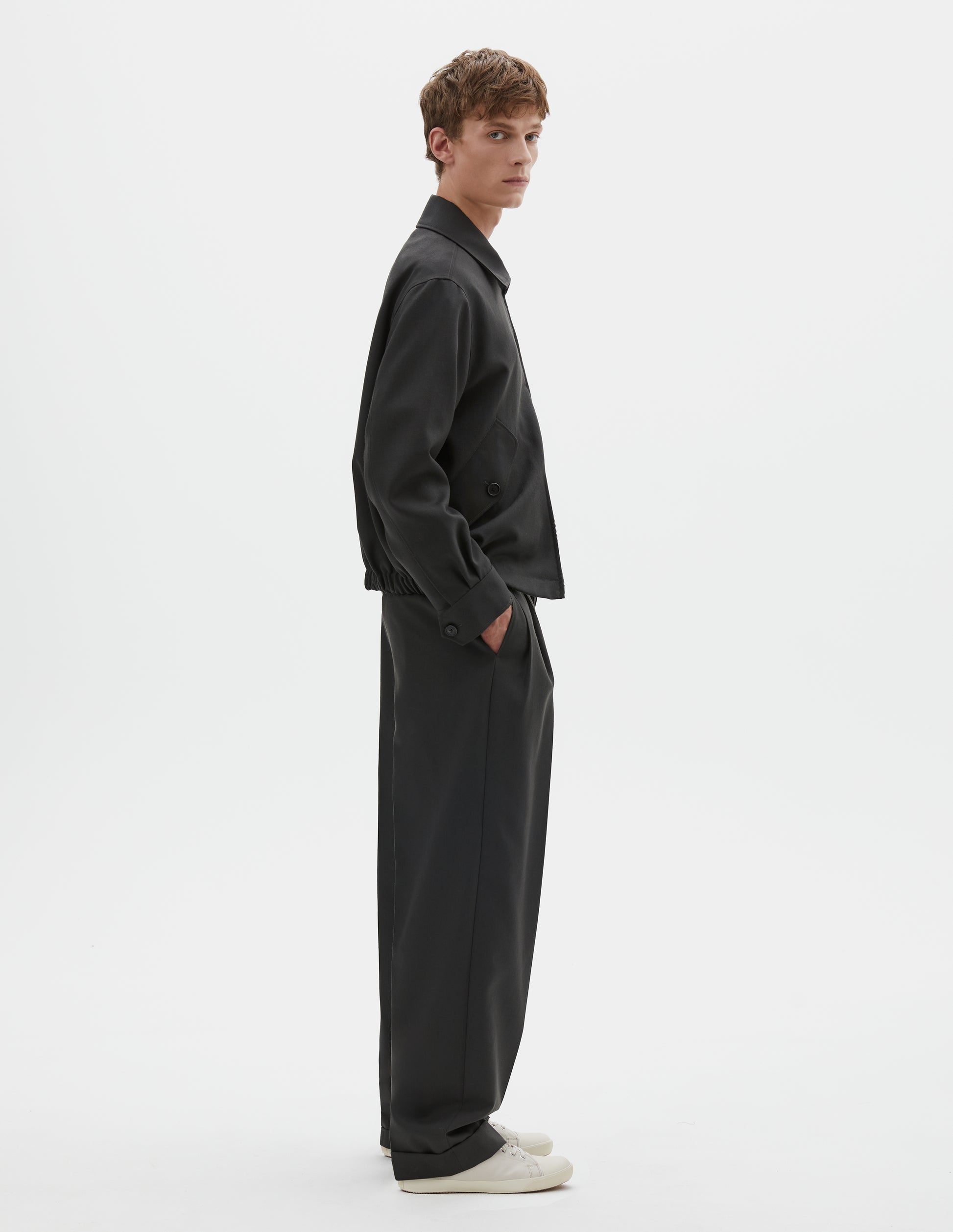Full body image, side view of model wearing dark grey wool jacket with two way zip fastening, elasticated waistband, tab shirt cuffs and button flap pockets. Styled with black pleated trousers and white canvas trainers.