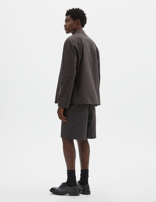 Full body image, back view of model wearing relaxed greyish brown blazer. Styled over green t-shirt, matching grey shorts and black socks with black leather shoes.