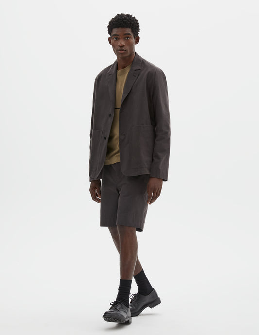 Full body image, front view of model wearing relaxed greyish brown blazer with horn button fastening and patch pockets. Styled over green t-shirt, matching grey shorts and black socks with black leather shoes.