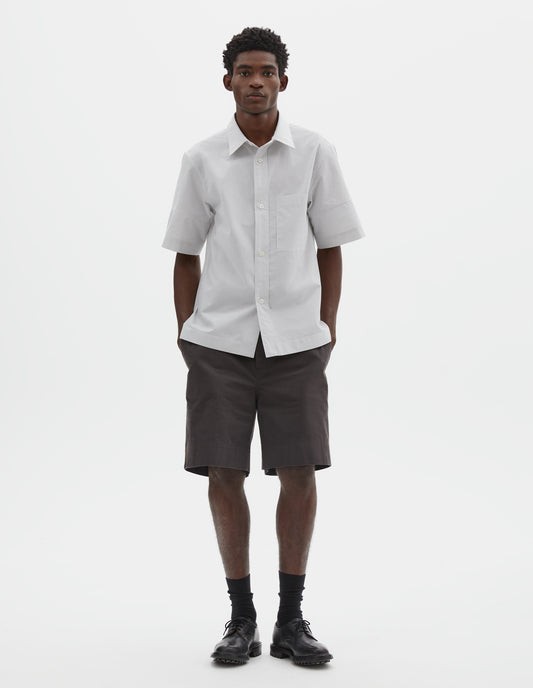 Full body image, front view of model wearing white shirt with grey stripes, panels on short sleeves with contrasting band at cuff, mother of pearl buttons on raised placket, bellows chest patch pocket and side split vents. Styled with dark brown shorts, black socks and black leather shoes.